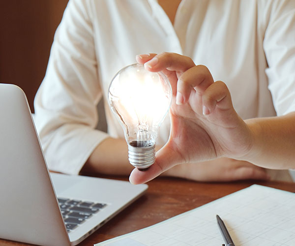 Bilden visar en kvinna framför sin bärbara dator som håller i en lysande glödlampa